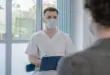 Ein Arzt in heller Arbeitskleidung spricht mit einer Patientin oder einem Patienten und macht Notizen.