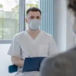 Ein Arzt in heller Arbeitskleidung spricht mit einer Patientin oder einem Patienten und macht Notizen.