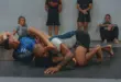 Zwei Männer beim Grapplingtraining auf einer Matte; im Hintergrund sind weitere Sportler zu sehen.