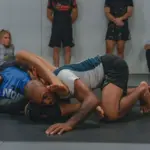 Zwei Männer beim Grapplingtraining auf einer Matte; im Hintergrund sind weitere Sportler zu sehen.
