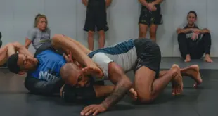 Zwei Männer beim Grapplingtraining auf einer Matte; im Hintergrund sind weitere Sportler zu sehen.