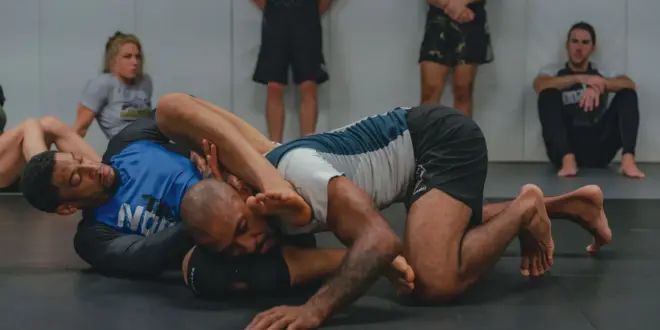 Zwei Männer beim Grapplingtraining auf einer Matte; im Hintergrund sind weitere Sportler zu sehen.