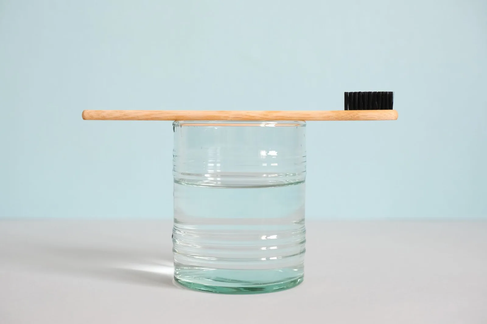 Eine Bambuszahnbürste liegt waagerecht auf einem halb gefüllten Wasserglas vor hellblauem Hintergrund.